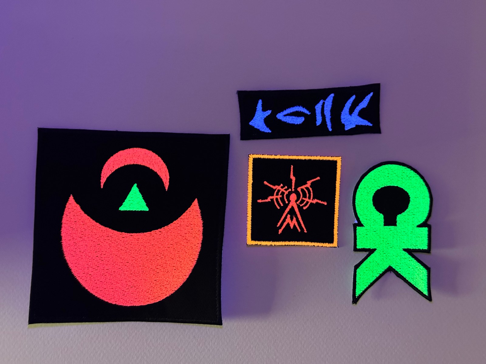 Four custom fluorescent embroidered patches glowing under UV light: a red crescent design, a blue alien-script strip, an orange-bordered square, and a bright green symbol